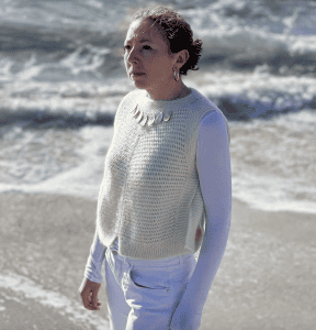 Frau trägt weiße gestrickte Weste am Strand, Meereswellen im Hintergrund.