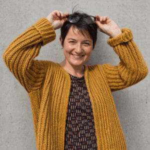 Frau trägt den Cardigan KABOCHA von DNK, lächelnd, vor einer grauen Wand.