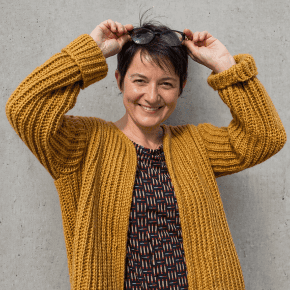 Frau trägt den Cardigan KABOCHA von DNK, lächelnd, vor einer grauen Wand.
