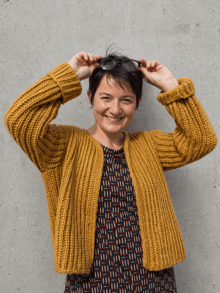 Frau trägt den Cardigan KABOCHA von DNK, lächelnd, vor einer grauen Wand.