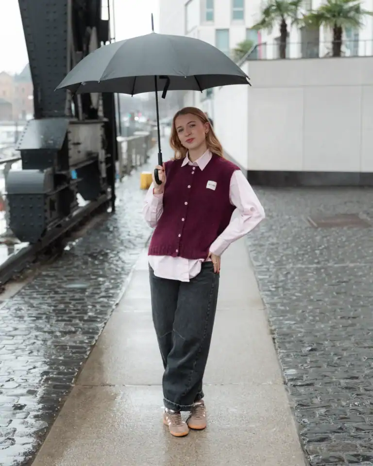 Frau mit Regenschirm auf nasser Straße, trägt lässigen Pullover und Jeans, bei regnerischem Wetter i.