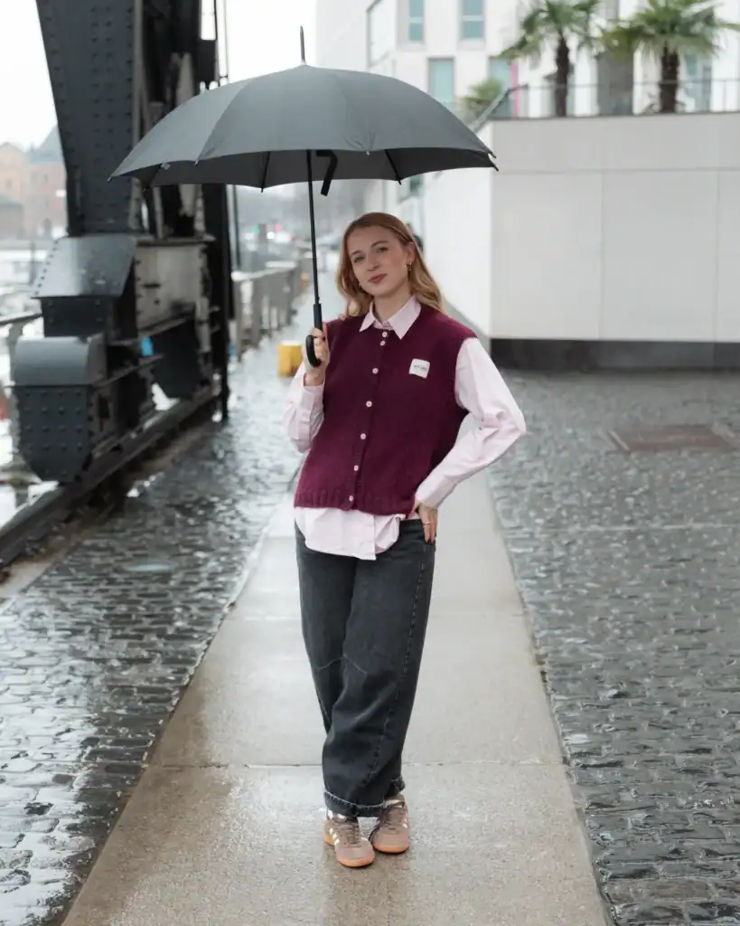 Frau mit Regenschirm auf nasser Straße, trägt lässigen Pullover und Jeans, bei regnerischem Wetter i.